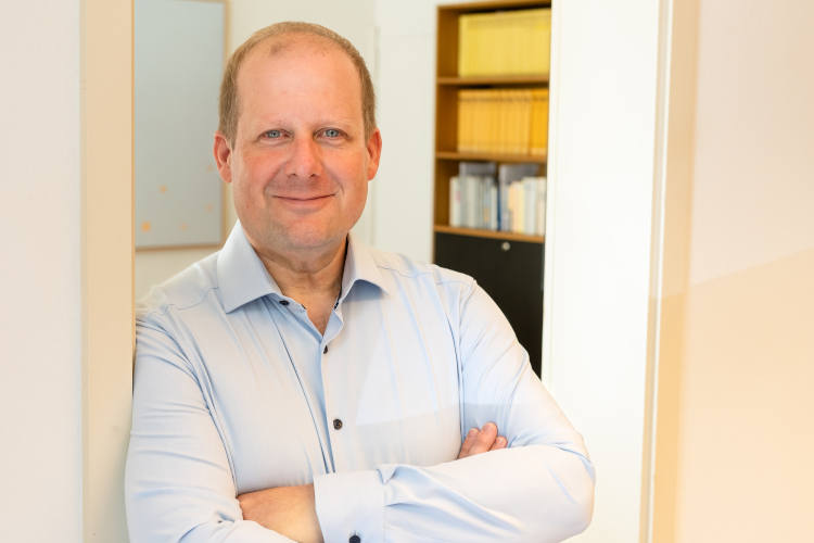 Ein Mann mit blondem Haar und blauem Hemd steht lächelnd mit verschränkten Armen in einem Büro, im Hintergrund ist ein Bücherregal zu sehen.
